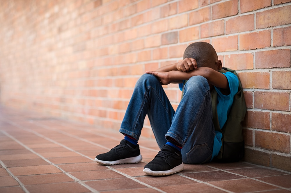 Young boy sitting alone with sad feeling at school. Depressed african child abandoned in a corridor and leaning against brick wall. Bullying, discrimination and racism concept at school with copy space.